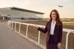 Erin Calder, l’autrice du guide, se tient à la rambarde d’un hippodrome et montre le chemin de la main ouverte, pendant qu’un avion monte dans le ciel du soir. L’image est générée par IA : c’est un personnage, et la piste n’est pas Century Mile.